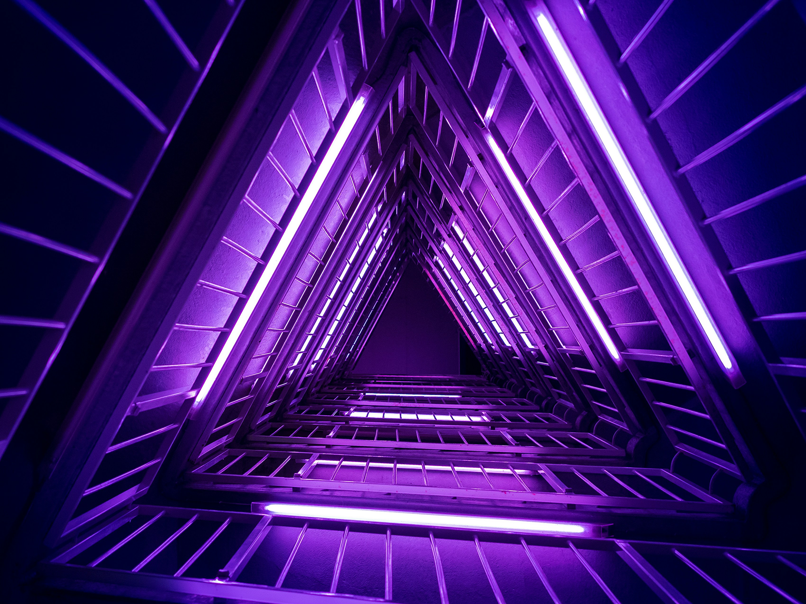 Looking up a stairwell of nested triangular landings lit by purple fluorescent tubes